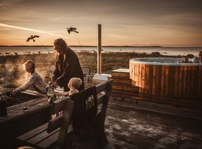 Mamma och två barn som står och grillar på ett trädäck. I närheten av dem syns en badtunna i bakgrunden som också är i trä, där resten av familjen badar.