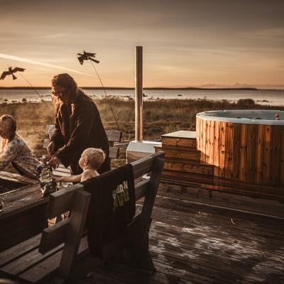 Sommarkväll på trädäck Mamma och två barn som står och grillar på ett trädäck. I närheten av dem syns en badtunna i bakgrunden som också är i trä, där resten av familjen badar.
